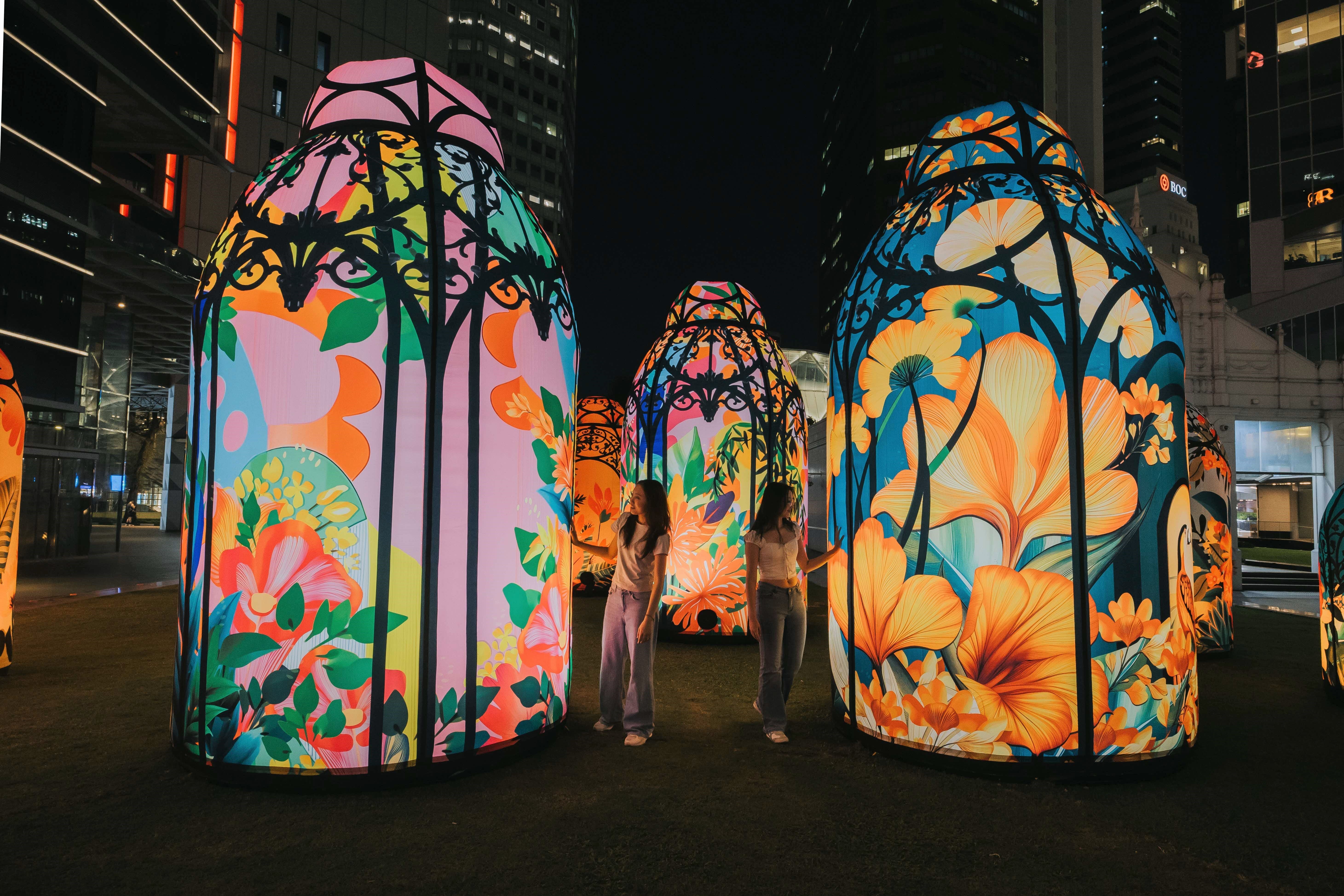 Two women standing among illuminated floral light installations at night in a city park.